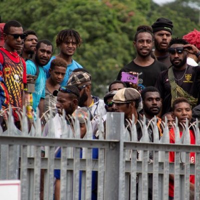 A group of about 16 men from PNG stand watching something over a fence, some with cameras